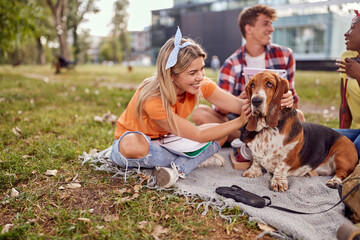A cute dog enjoys being hugged by young girl sitting on the grass with her friends in the park. Friendship, rest, pets, picnic