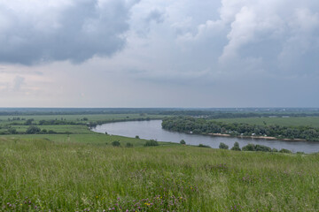 Summer landscape with meadow grass in the foreground. Dark sky with clouds before rain. Bright green grass. Wide river in the background.