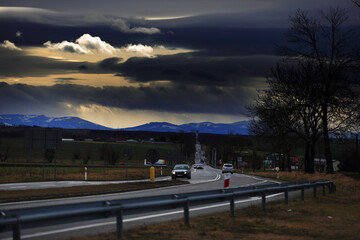 Samochody na drodze szybkiego ruchu, krajobraz wiejski przed zachodem słońca, góry i ciemne chmury.	