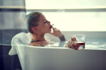 A young sexy tattooed girl with a cigar and a drink is having a bath in the bathroom. Bathroom, relaxation