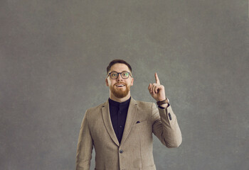 Studio portrait happy emotional handsome bearded young man in smart jacket and stylish geek glasses pointing index finger up. Entrepreneur or businessman astonished by clever brilliant creative idea