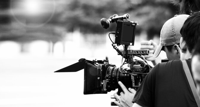 Back angle view of high definition digital video camera which recording or shooting tv commercial production on pan tilt shift professional head tripod at outdoor location and film crew team with set.