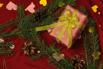 A little red gift box with gold ribbon among spruce branches and pinecone on red silk background