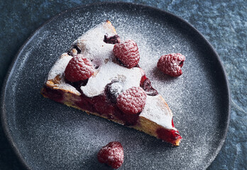 Raspberry cake garnished with fresh raspberries on dark plate with powder sugar. Flat lay of homemade raspberry cake dessert on blue plate. Raspberry cake dessert.