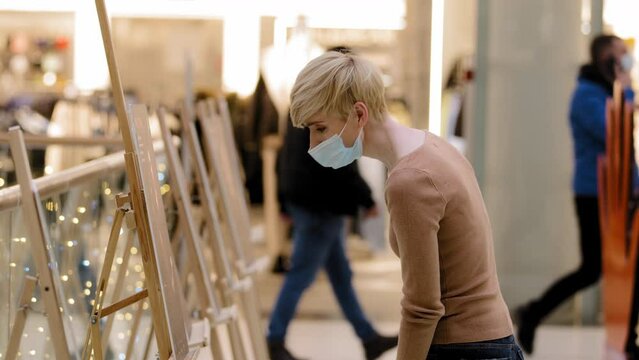 Side view caucasian middle aged woman lady in medical mask art female artist in public place at exhibition of paintings looks at portraits landscapes drawings on easels visits exhibition contemplates