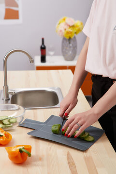 Shot of a woman cutting a cucumber on a black plastic cutting board. There are bell peppers on a table, cucumber slices in a glass bowl. There is a sink, a wine bottle and flowers on the background.