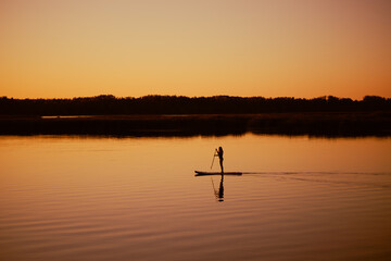 Fototapeta premium Silhouette of woman doing sup boarding alone on lake at sunset time holding oar in hands with charming yellow orange sky, water and trees in background. Active lifestyle.