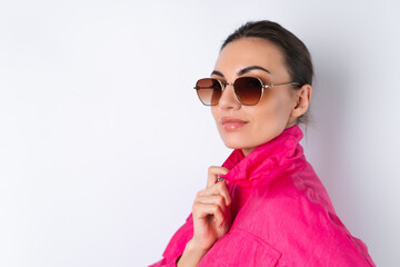 Stylish young woman in a bright pink sports jacket, trendy sunglasses on a white background looks at the camera, side view