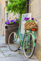 Fototapeta na wymiar Finalborgo, Finale Ligure, Italy. May 5, 2021. In a small street in the center an old green bicycle leaning against a wall with wicker baskets with flower pots inside.