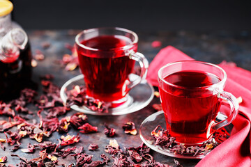 Hibiscus tea in glass cup and a burgundy napkin. Cup of hibiscus tea and dry hibiscus petals on a dark background. Healthy natural slimming drink
