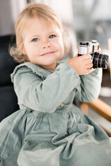 Portrait of adorable cute baby girl holding in arms little photo camera, smiling, wearing stylish green dress. Beautiful lovely toddler sitting on chair and looking at the camera