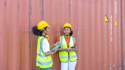 Two African female sisters in safety uniform and hardhat use computer to have a conversation next to a blue shipping container at a cargo storage. Female supervisor provides instruction to trainee