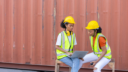 Two African female sisters in safety uniform and hardhat use computer to have a conversation next to a red shipping container at a cargo storage. Female supervisor provides instruction to trainee