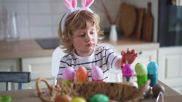 Cute two-year-old boy in bunny ears paint Easter eggs with multi-color paints sitting at the table on the kitchen Video