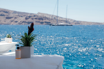 Fototapeta na wymiar Blurred sea view with a yacht from a restaurant with white tables, flowers on the table and alcohol gel, Santorini island, Oia, Greece. Vacation during the coronavirus epidemic. Nice seascape