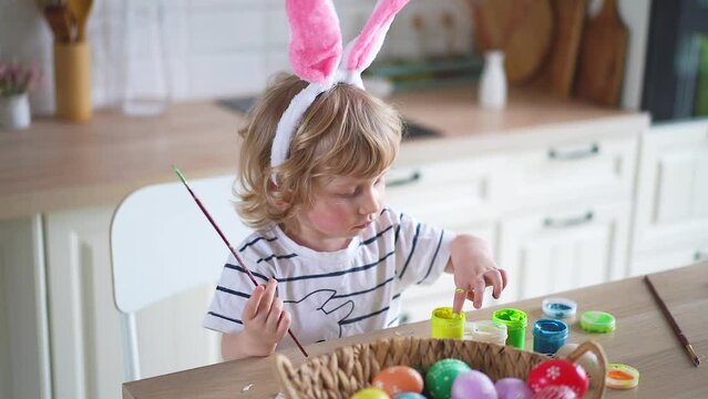 Cute two-year-old boy in bunny ears paint Easter eggs with multi-color paints sitting at the table on the kitchen Video