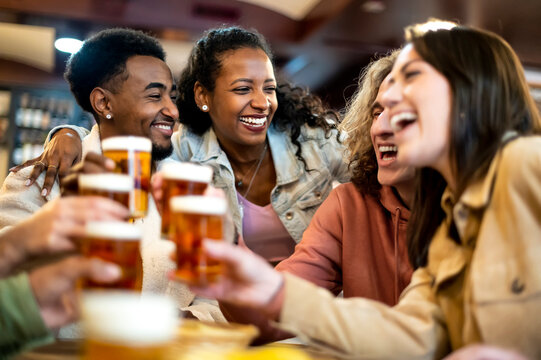 Group of smiling friends drinking and toasting beer at bar restaurant - Friendship concept with young happy people having fun together toasting brew pint on happy hour.