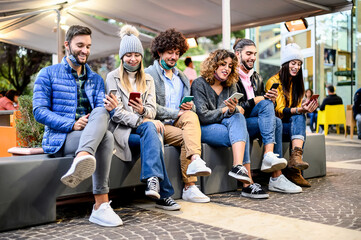 Young happy friends with face mask down sitting on bench using smartphone watching social media content online outside the bar - Concept of youth, tech and friendship - People Working Together