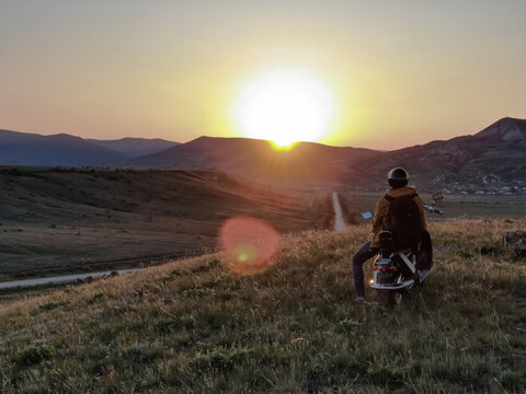 A man with a backpack sits on a scooter against the backdrop of a mountain range and admires the sunset