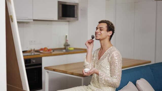 A transgender guy in fancy dress doing make up with brush and powder