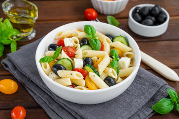 Healthy pasta salad with tomatoes, cucumbers, red onions, olives and cheese in a bowl on a dark wooden background. Copy space.