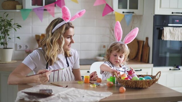 charming woman and her two-year-old son in bunny ears paint Easter eggs with paints sitting at the table on the kitchen Video HD