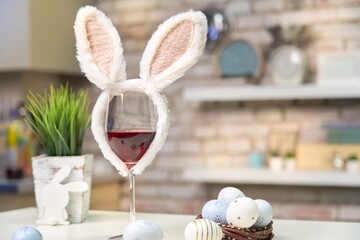 Glass of rose or red wine with bunny ears and Easter decorations, colorful eggs on white table in the kitchen at home. Copy space for text.