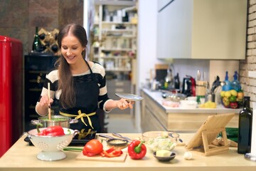 Happy young woman cooking in kitchen, wearing apron.