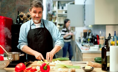 Handsome older man wearing apron, cooking at home in the kitchen, cutting vegetables on board.