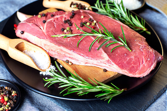 Raw beef steak on a black plate and wooden spoon on spices and rosemaryon grey concrete background. Top view close up.