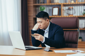 Upset Asian man, trying to make an online purchase, sitting in the office, uses a credit card, and a laptop, trying to remember the pincode