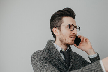 a young stylish man with a beard and glasses in a shirt and a gray cardigan against a gray wall is...