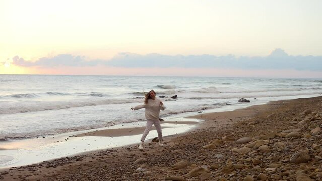 Happy young woman with long blonde hair in white suit running along ocean beach at sunset