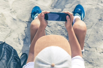 View from the top of young boy watching video in the street sitting on the ground. Hipster male enjoying podcast outdoors with backpack out of school Teenager casual dressed reads news with smartphone