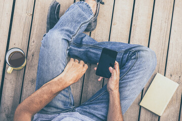 Top view of a boy using smartphone sitting on a wooden floor. Cup of coffee and book on the side....