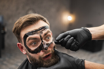 Frightened man grimaces while removing black facail skin mask in beauty salon