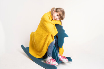 A 7-8-year-old girl with dark hair is covered with a warm yellow knitted plaid, sitting on a blue wooden rocking horse. Accessories for sleeping and home comfort in the children's room.