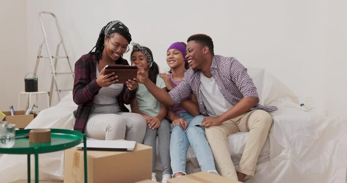 Smiling couple with two daughters are sitting on sofa in renovated apartment woman is holding a tablet in hands, taking a selfie with her loved ones, laughing, joking, failed photo