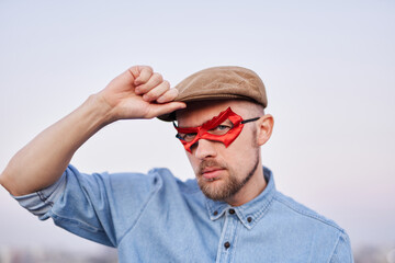 Cute man in red superhero mask and jeans shirt posing in front of the camera correcting peaked cap outdoor at sunset with urban view. Man power concept. High quality image