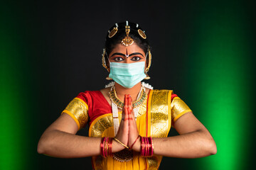 Portrait shot of Bharatnatyam dancer with medical face mask greeting by doing namaste while looking at camera - concpet of protection against covid-19 coronavirus infection and pollution
