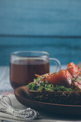 Bruschetta with salmon and avocado on a wooden plate on a blue wooden background on a white wooden table with a cup of tea.