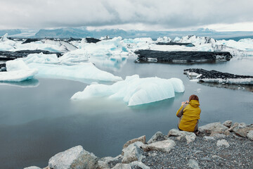 A person sitting by Jökulsárlón Glacial Lagoon, taking a photo with a small camera. Icebergs on...