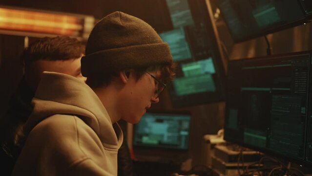 Young man hacker in hat and glasses talking with associate and using computers to hack data base while sitting at desk on dim criminal base