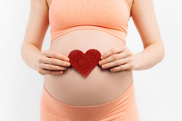 Young pregnant woman holding and touching her belly, holding a red heart sign, belly close-up