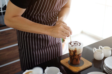 Asian barista young man pouring fresh Chocolate art in a plastic glass of Cappuccino iced coffee for according to order at counter bar in the morning at modern cafe.