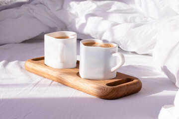 Coffee in mugs in the shape of a heart on a tray lie on a white bed. In the morning, the sun's rays illuminate the cups and a crumpled white sheet.