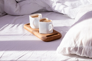 Fototapeta na wymiar Coffee in mugs in the shape of a heart on a tray lie on a white bed. In the morning, the sun's rays illuminate the cups and a crumpled white sheet.