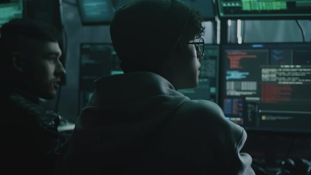 Young male hacker in glasses looking at screen and talking with associate while performing cyber attack on computer in dim room of criminal base