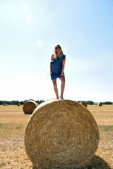 Naklejka na ściany i meble Young woman in sunglasses smiling stands on a round haystack in a cereal field