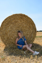 Naklejka na ściany i meble A young Caucasian woman in a blue dress sits near a round haystack in a cereal field
